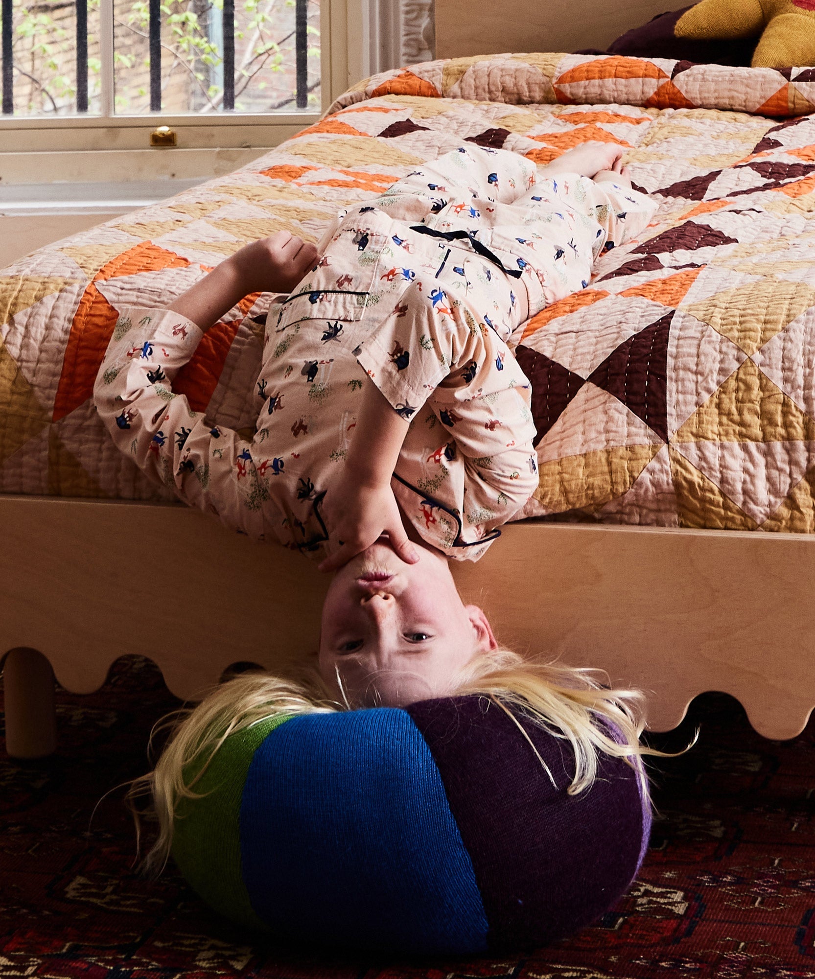 Child in pajamas lies upside down on a bed, head resting on the Circus Ball - Rainbow cushion, in a cozy room.