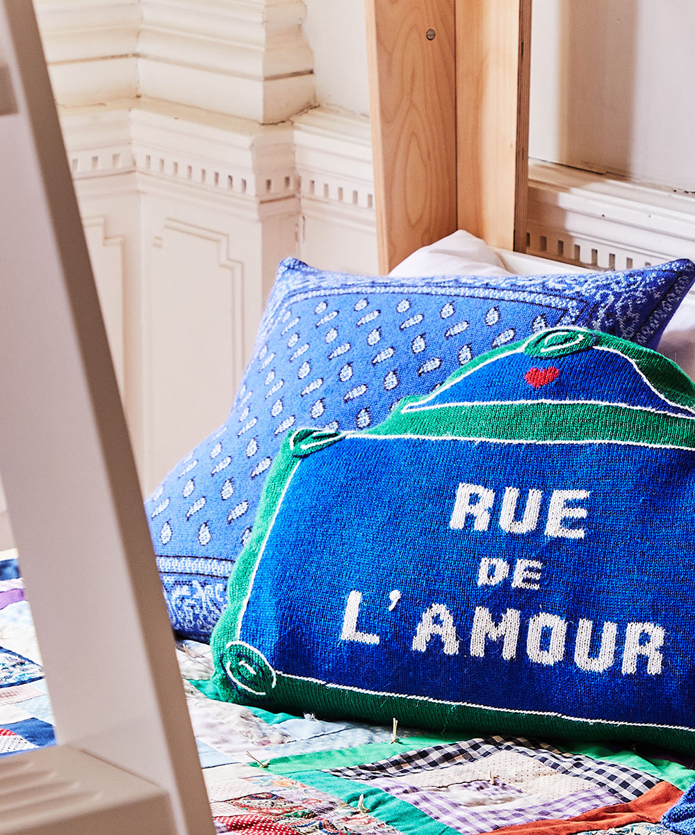 A cozy room features a bed adorned with patterned pillows, including a blue "Rue de L'Amour" and Bandana Pillows.