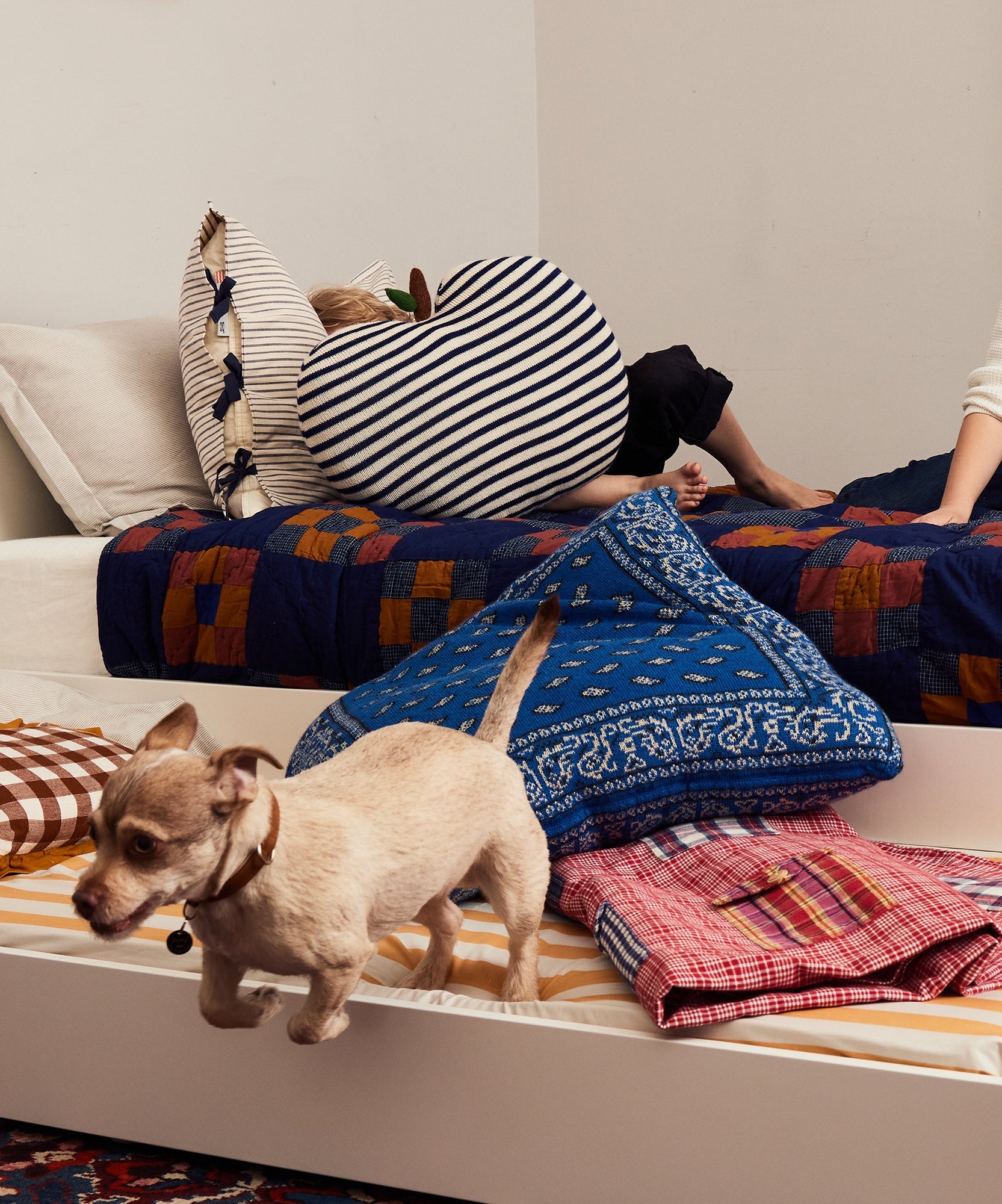 A small dog jumps off a bed while a child holding a Bandana Pillow sits on another bed.