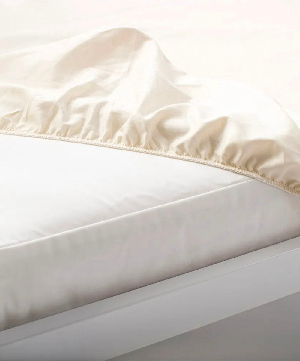 A close-up of the Organic Waterproof Crib Mattress Protector Pad in beige, partially covering a mattress on a white bed frame.