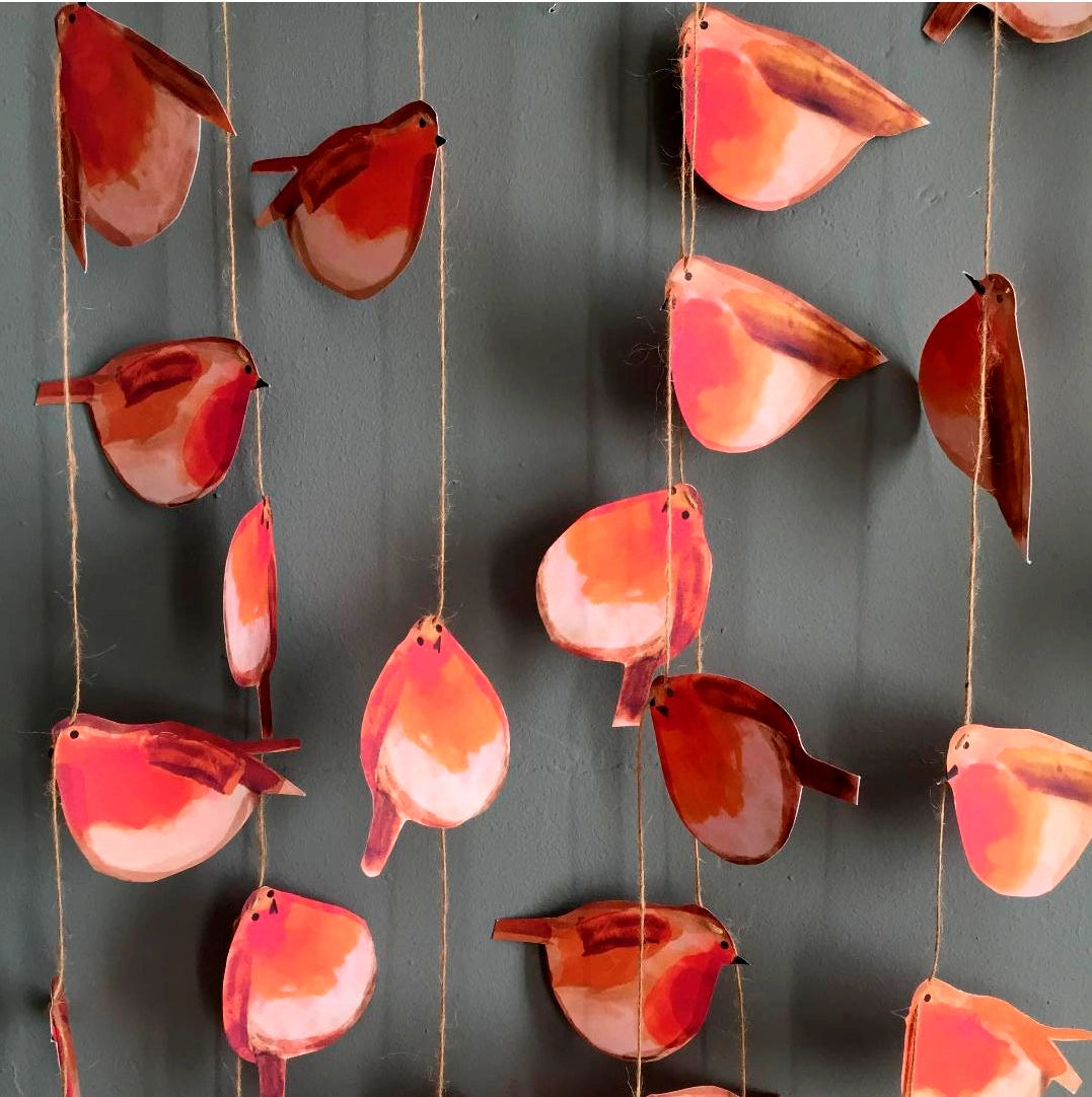 Le Garland - Robins features red and orange paper robin bird cutouts hanging on strings against a gray wall.