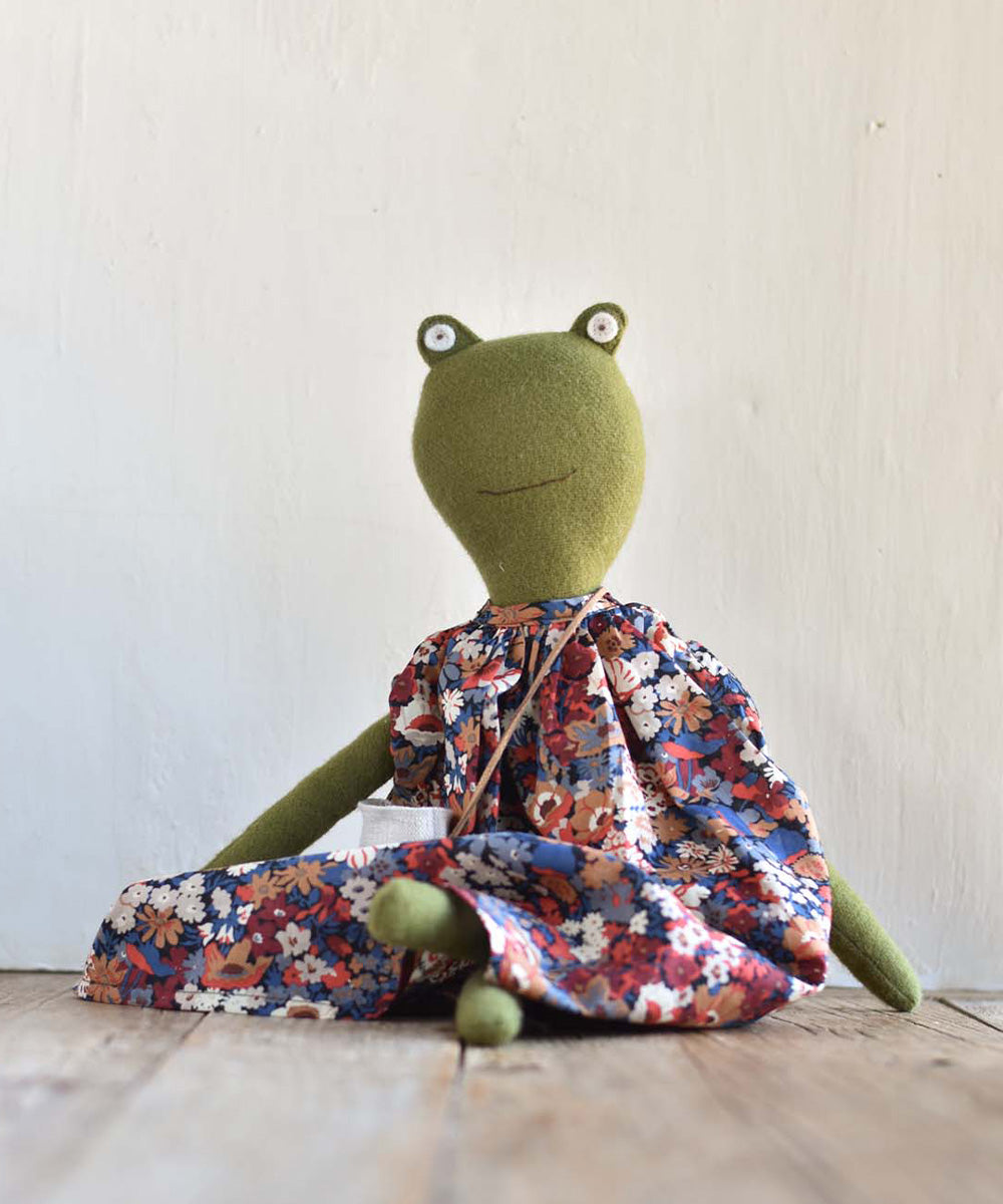 Fern le Frog – Canyon Liberty Dress: A green plush frog doll in a floral dress sits on a wooden floor by a light-colored wall.