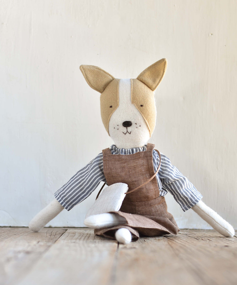 Fig le Frenchie plush dog wearing linen overalls, seated on a wooden floor against a plain wall.