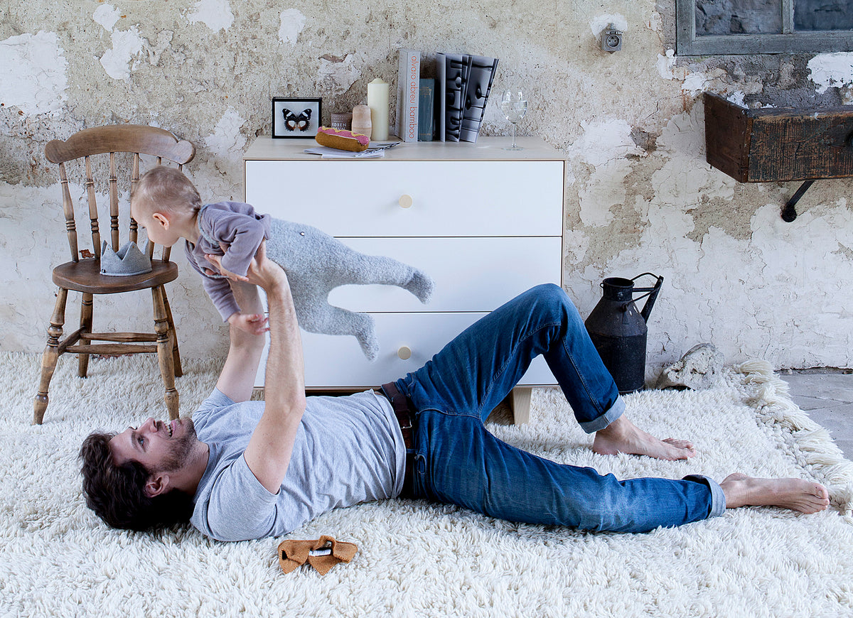 Man lying on a white rug lifts a baby in the air. A wooden chair, white dresser, and rustic decor are in the background.