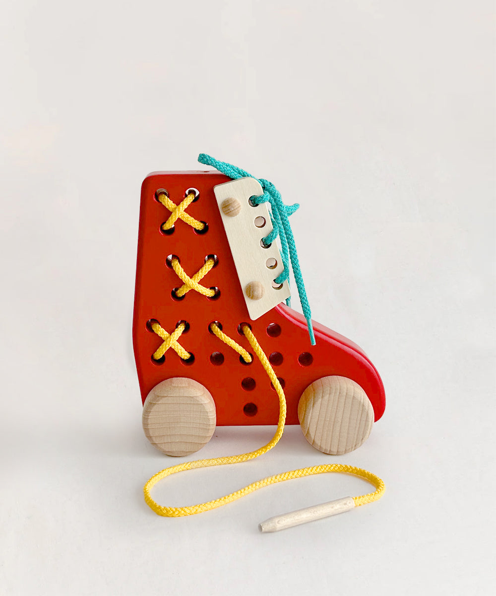 Le Lacing Shoe is a wooden toy with holes, yellow and blue laces, and wheels—perfect for practicing lacing and tying skills.