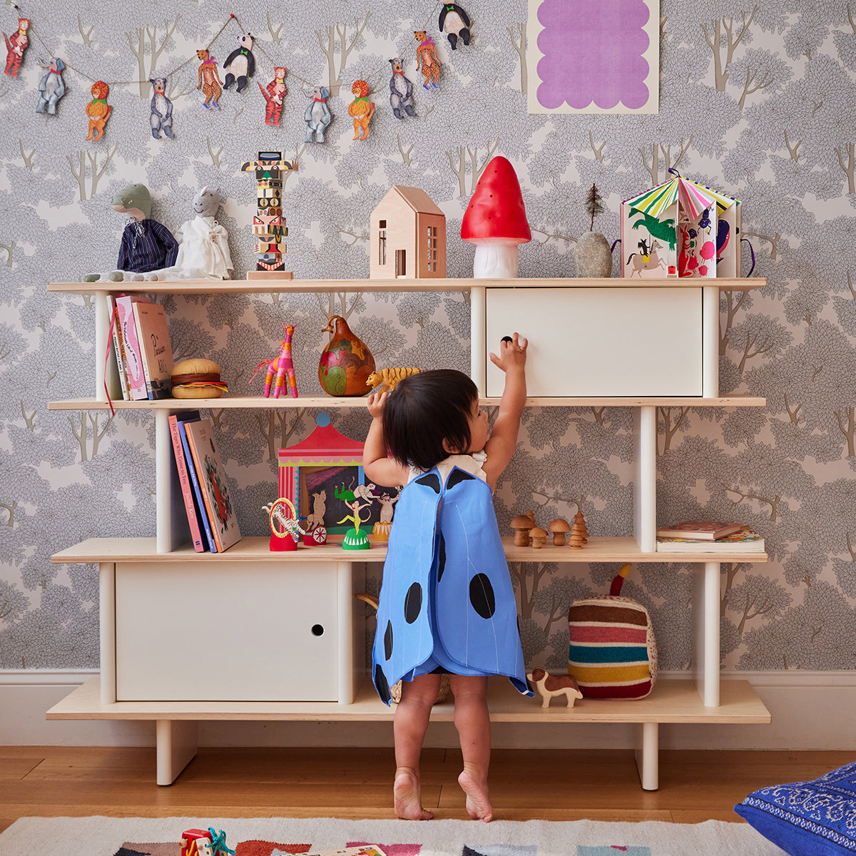 A child in a blue dress with black spots reaches up to a shelf filled with toys and ornaments in a playfully decorated room.
