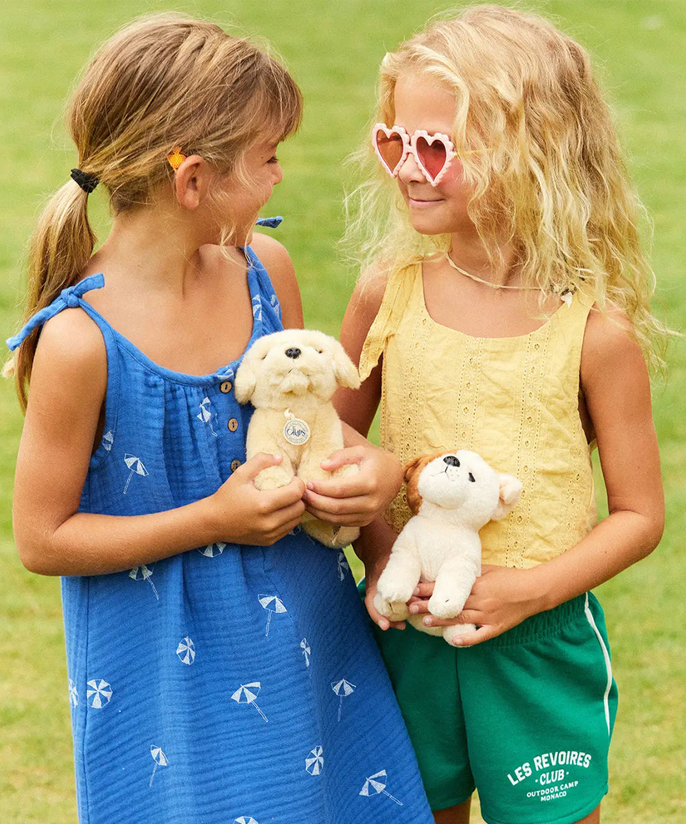 Two young girls smile on grass, holding Loulou le Labrador stuffed dogs. One wears a blue dress, the other wears yellow and green.