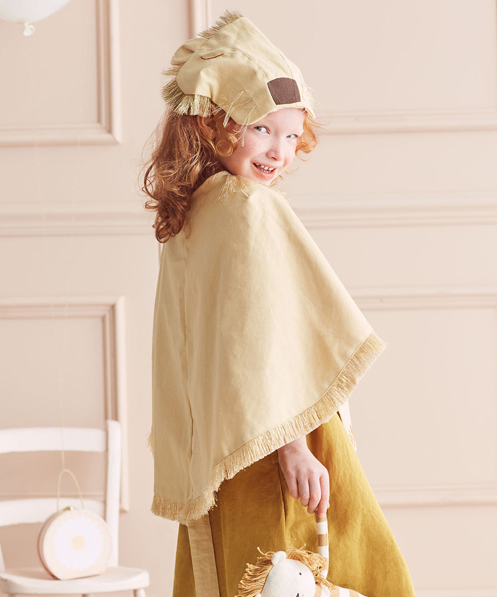 A child wearing the Lion Costume with a cape holds a plush lion toy while standing indoors.