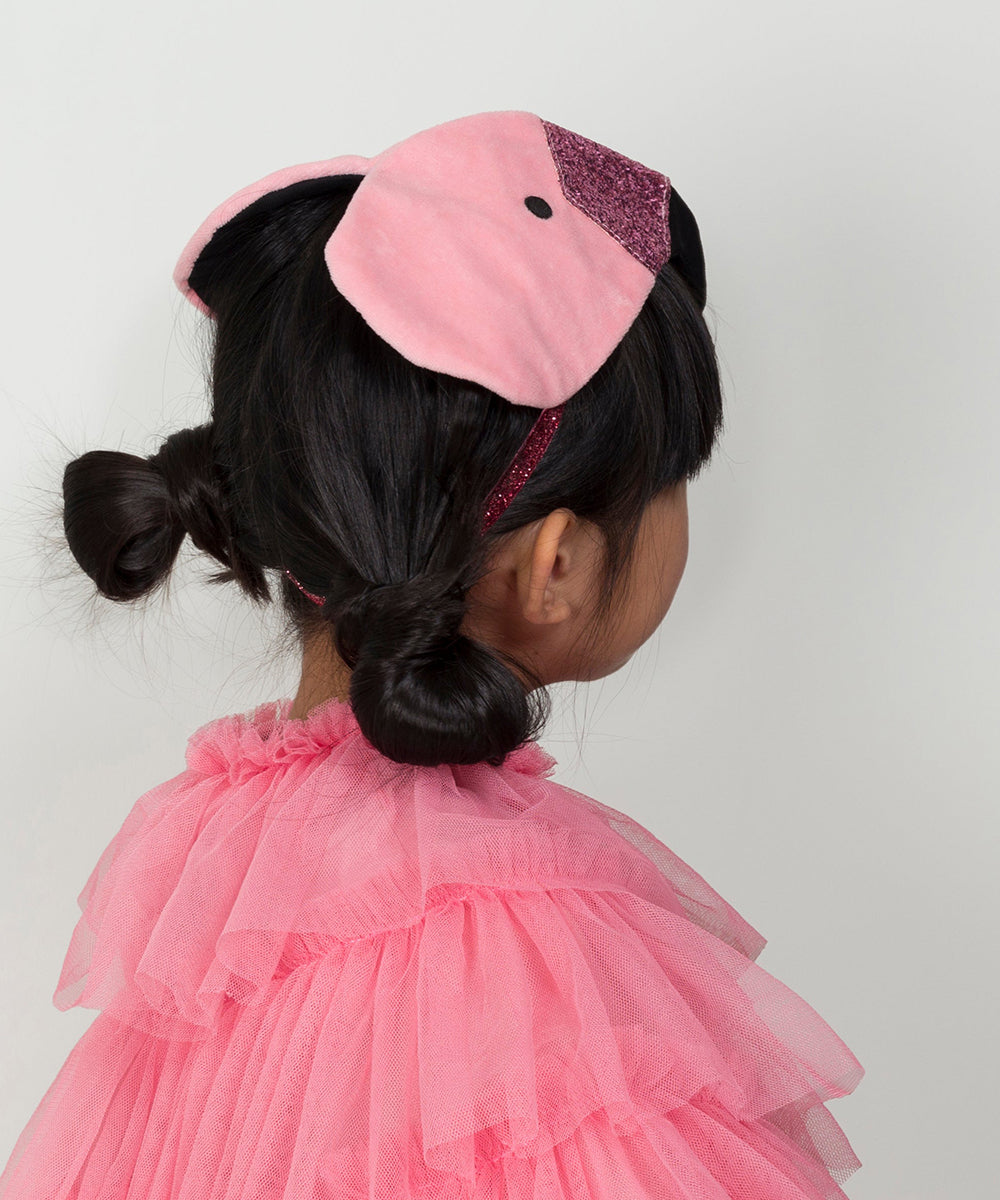 A child with dark hair in buns wears a Flamingo Costume, featuring a pink headband and ruffled dress, facing away.