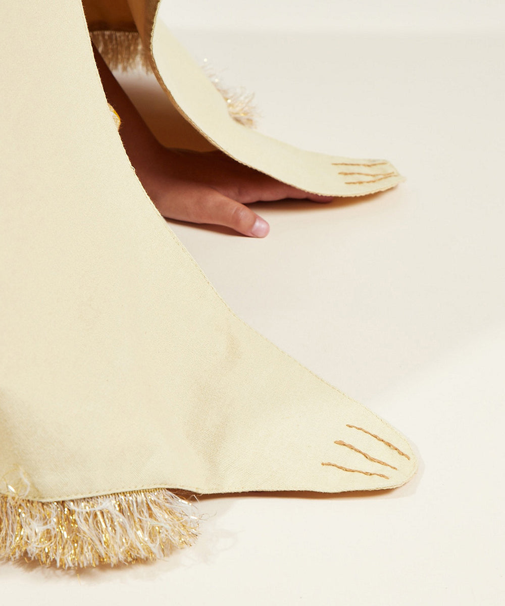 A hand touches the floor beside a cream-colored Lion Costume paw with gold fringe and stitching.