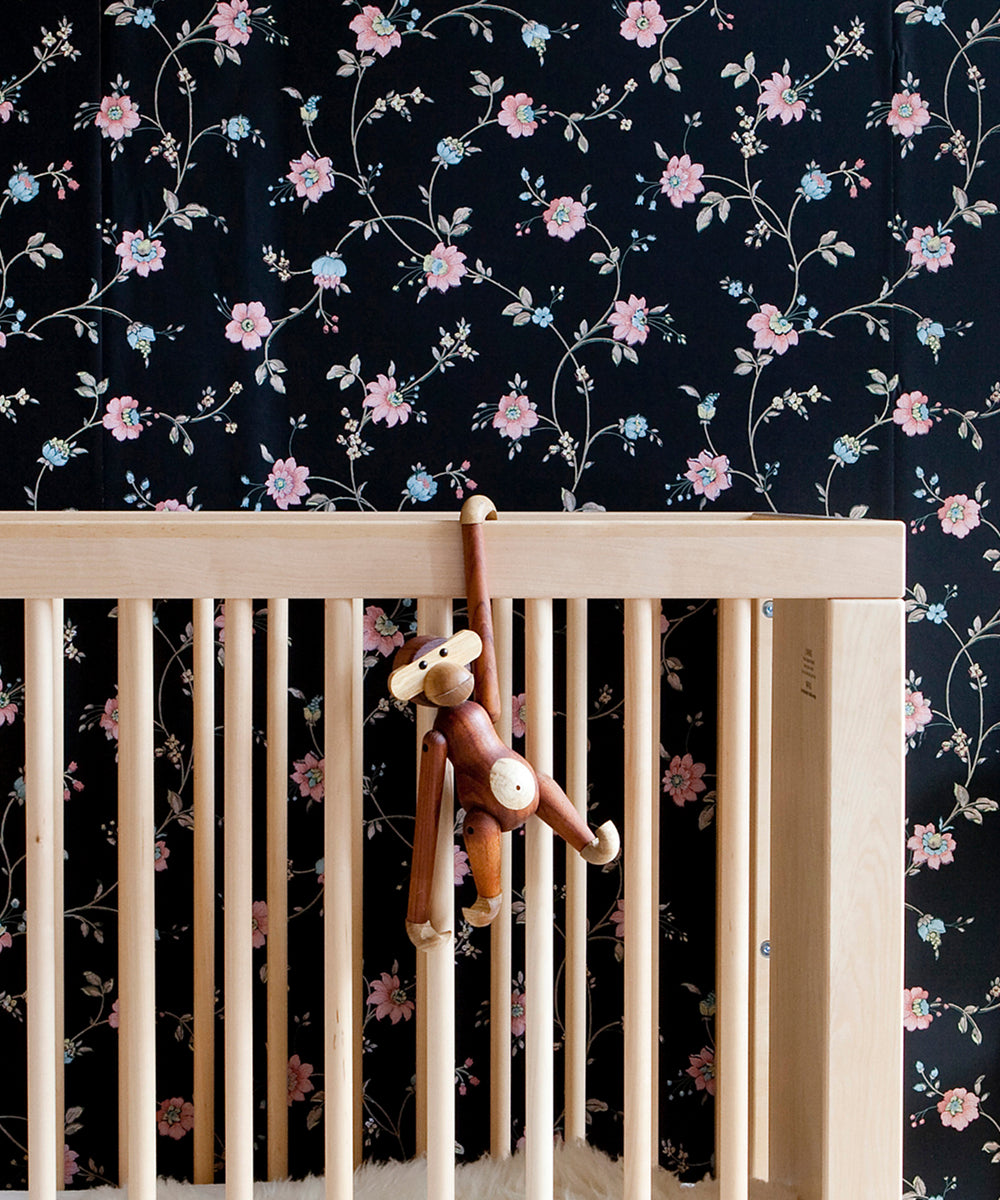 Le Monkey by Kay Bojesen hangs on a light wooden crib, set against a backdrop of black floral wallpaper.