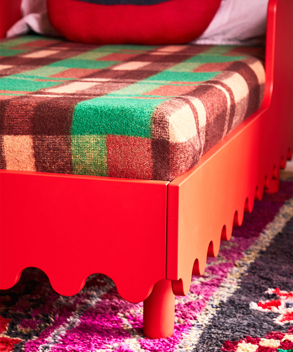 The Moss Toddler Bed features a red frame with scalloped edges, complemented by a green and brown plaid blanket, and sits atop a colorful, textured rug.