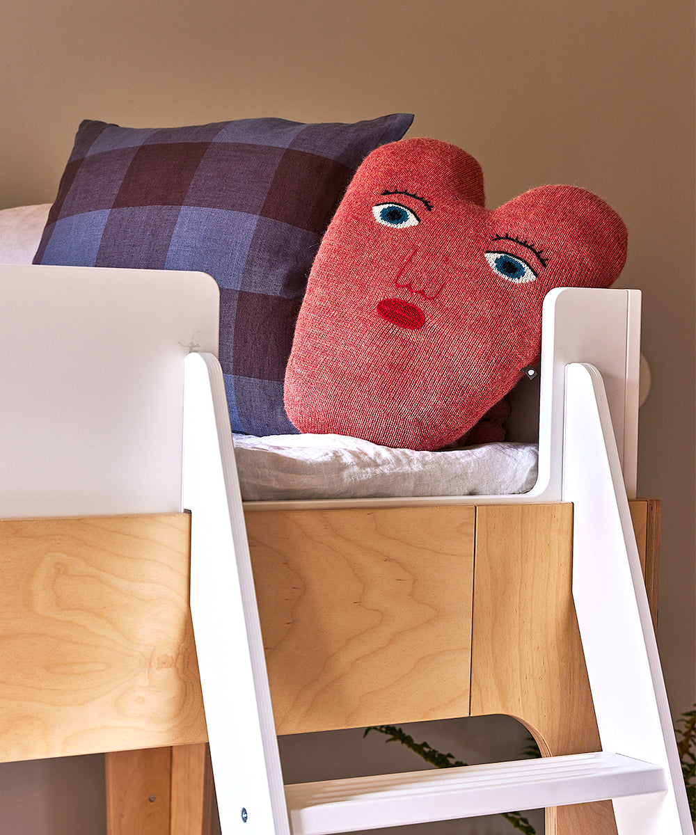 Heart Pillow with a face on a loft bed, beside a checkered pillow, with a white ladder in front.