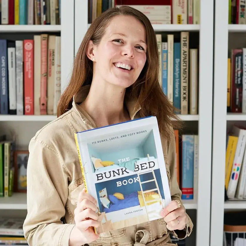 Person smiling and holding a book titled The Bunk Bed Book in front of a bookshelf filled with various books.