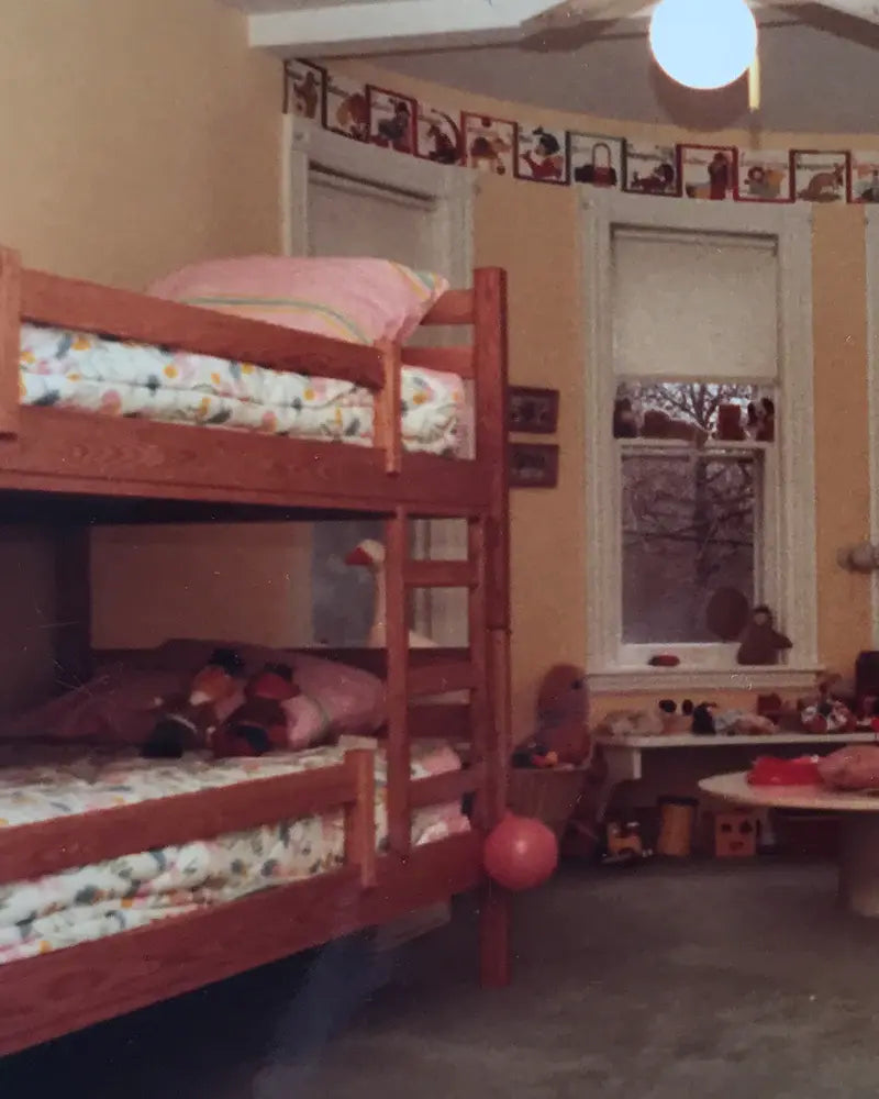 Kids room with wooden bunk beds, toys on the lower bed, and a small table with toys beneath two windows. A banner with hanging decorations spans the wall above the windows.