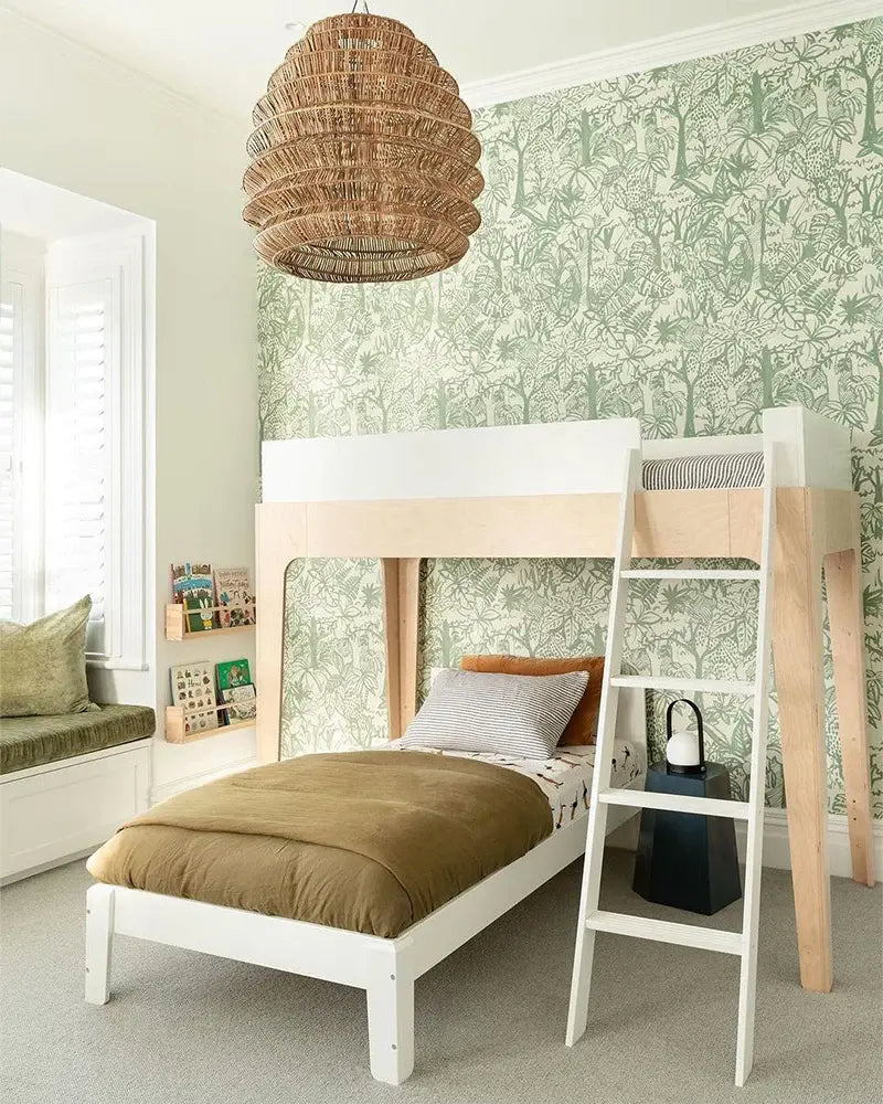 A bedroom with a loft bed, brown blanket, ladder, and light wood finish. Wallpaper with tree patterns, woven ceiling light fixture, and a window seat with cushions and books.