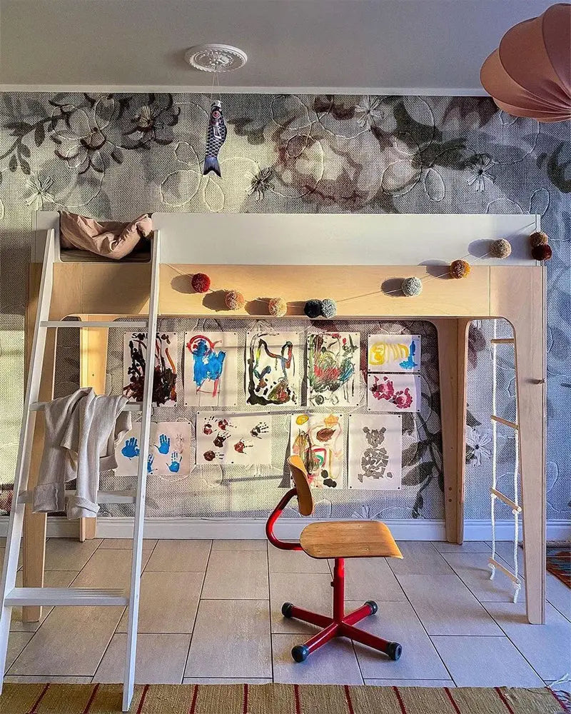 Loft bed with ladder and pom-pom garland, art display beneath on wall, red and wood chair on tiled floor, floral wallpaper background.