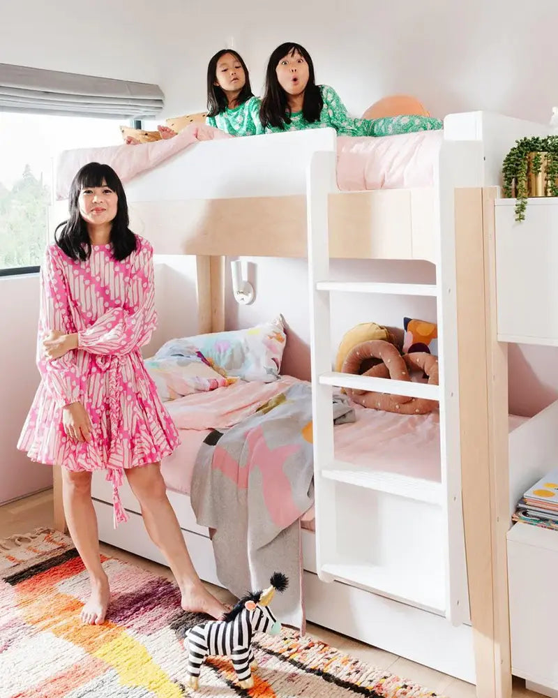 A woman in a pink dress stands by a bunk bed with two children sitting on the top bunk, in a colorful room with a striped rug and a toy zebra.