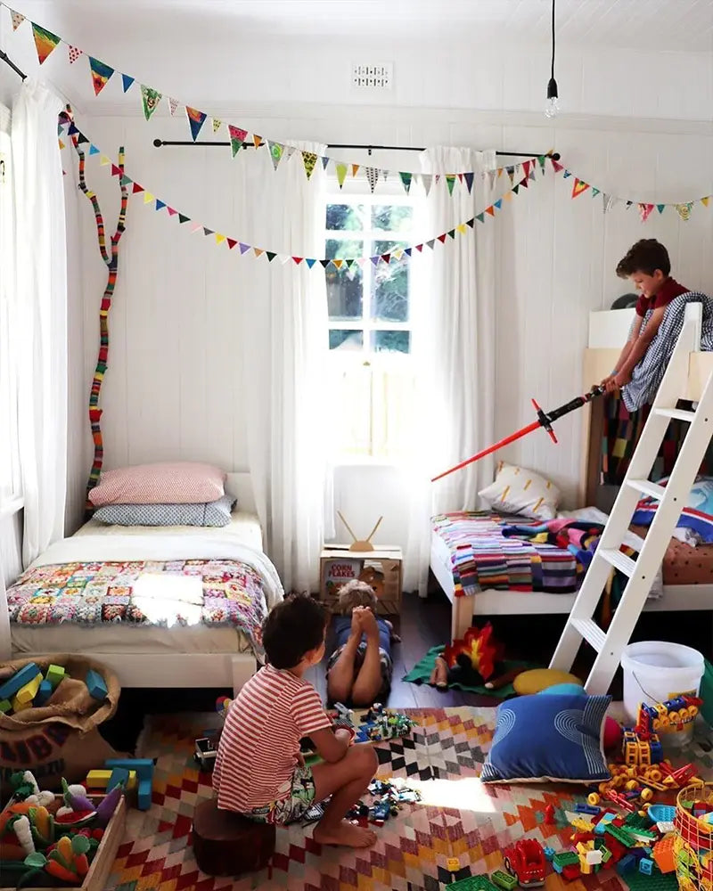 Children playing in a brightly decorated room with bunk beds, a tree mural, and colorful flags. One child plays with a toy sword on the top bunk, while others sit on the floor with toys.
