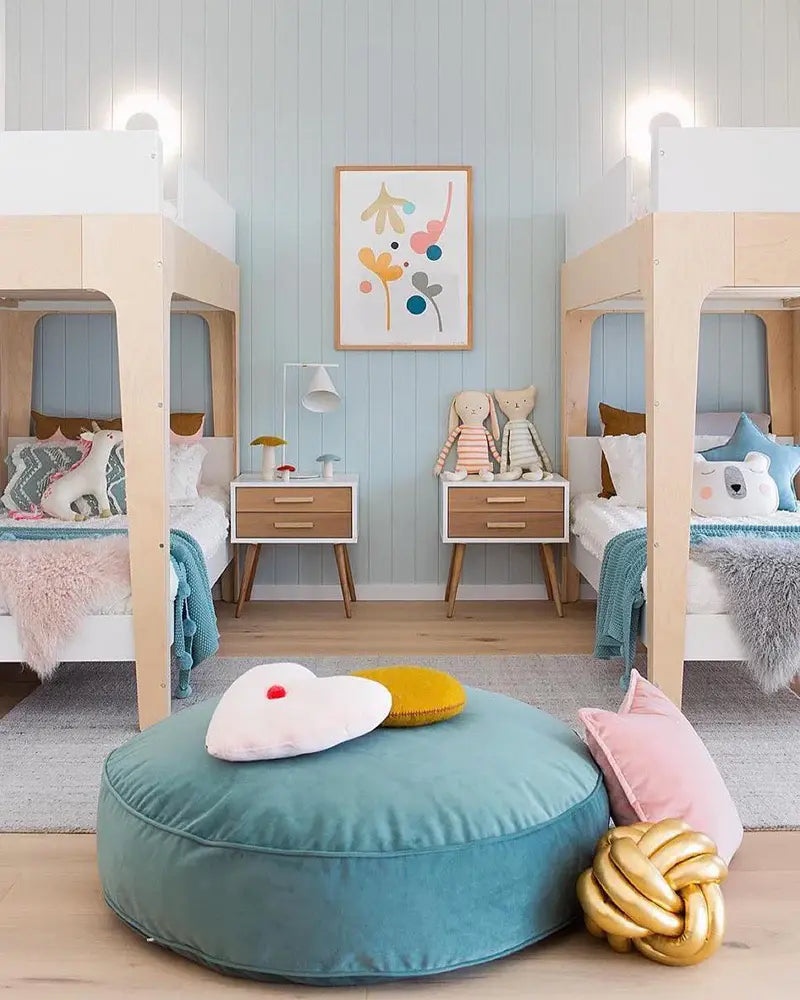 A childrens room with two bunk beds, colorful pillows, plush toys, a central teal cushion, and a framed abstract painting. The walls are blue with wooden accents.