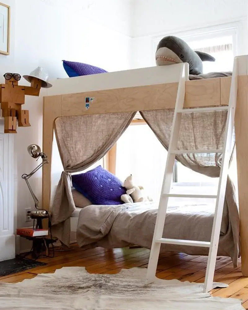 Childrens loft bed with a ladder, featuring a shark toy and purple pillow on top. A curtain encloses the lower bed, and a wooden floor is visible. Lamps and wooden figures decorate the room.