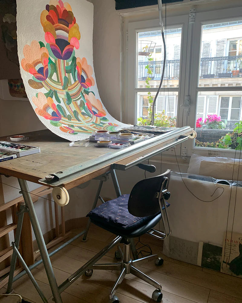 Brightly colored floral painting on a drafting table by a window, surrounded by art supplies and a chair.