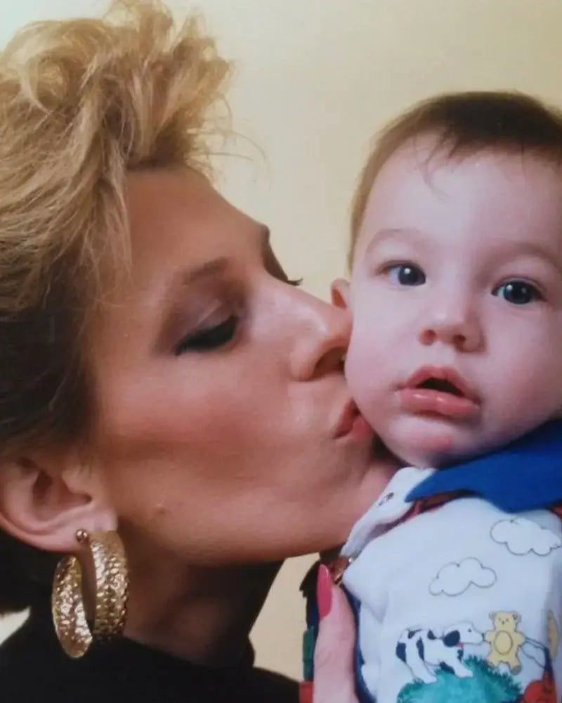 A woman with blonde hair kisses a baby on the cheek. The baby looks forward with a neutral expression. The woman wears large gold hoop earrings. Both have light skin tones.