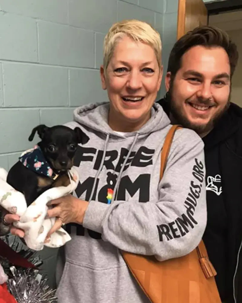 Two people pose happily with a small black and brown puppy wrapped in a blanket. The person on the left wears a gray hoodie with text, while the other wears a dark jacket.
