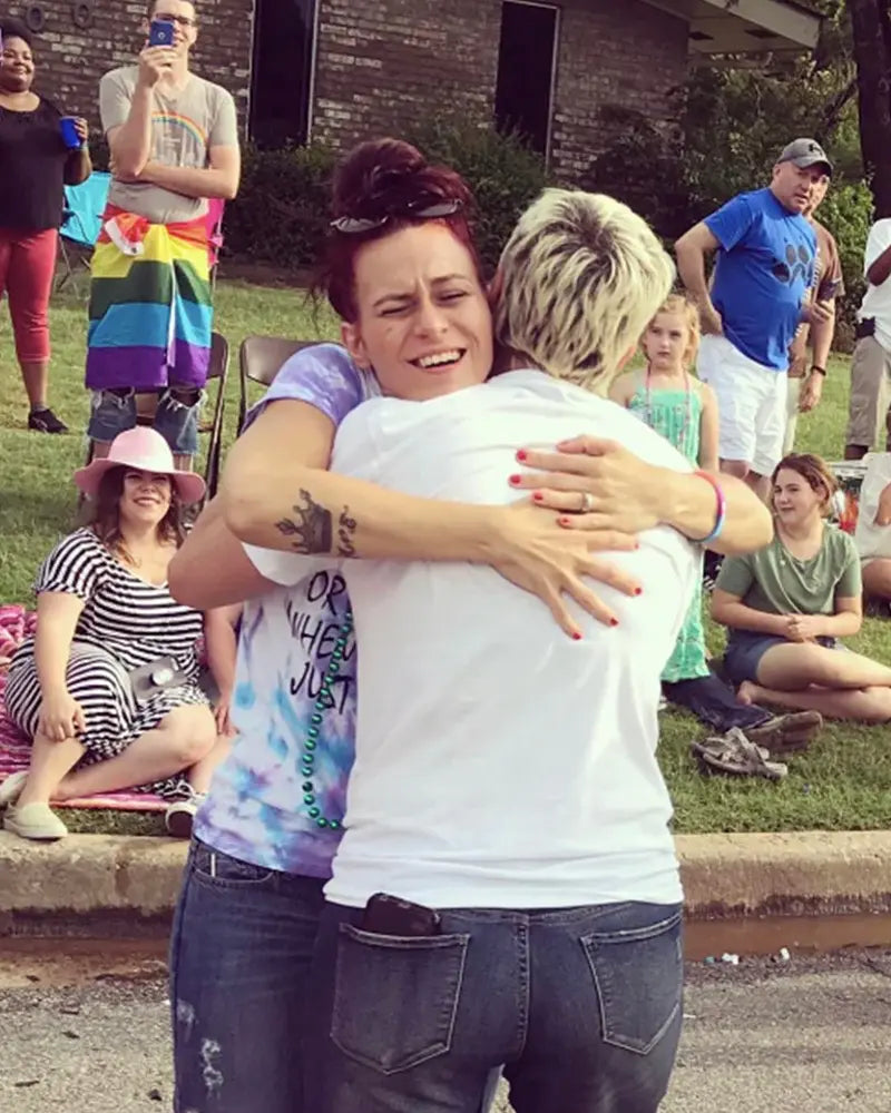 Two people hugging at an outdoor event with onlookers, including a person holding a rainbow flag. Grass and a building are in the background.