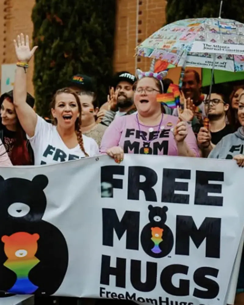 A group of people hold a Free Mom Hugs banner at a gathering. Some wear shirts with the same message, and one person holds a colorful umbrella.