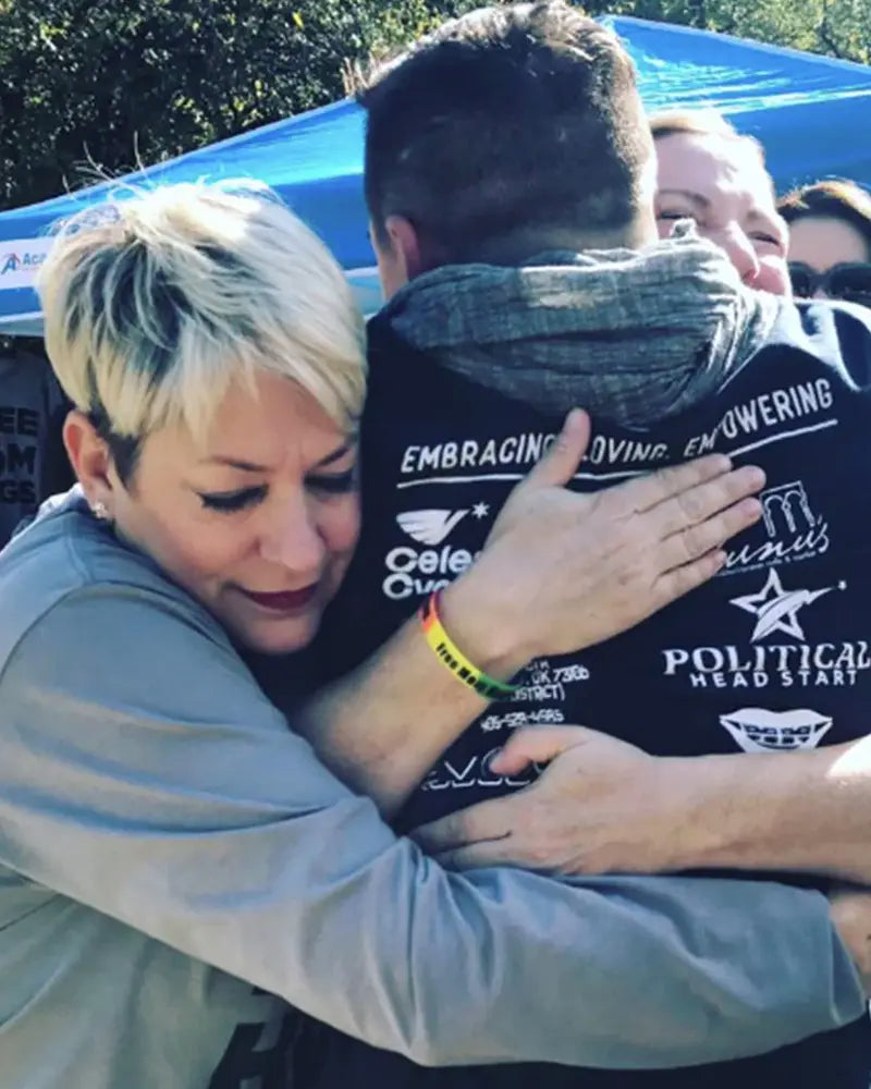 A person with short hair hugs another person wearing a jacket with logos and text, under a blue canopy.