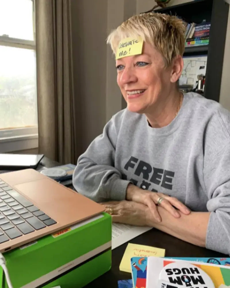 Person with a sticky note on forehead sits at a desk, smiling at a laptop. Books and a green box are on the desk.