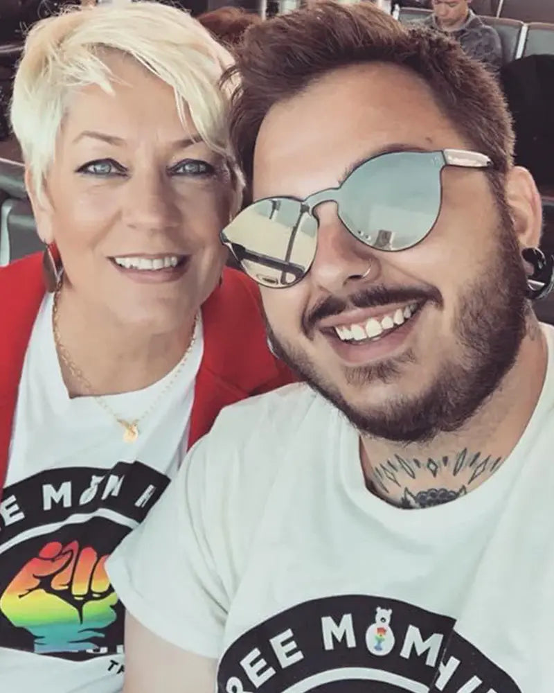 Two individuals are posing for a selfie. The person on the left has short, blonde hair and a red jacket. The person on the right has sunglasses, facial hair, and tattoos, wearing a similar white shirt.