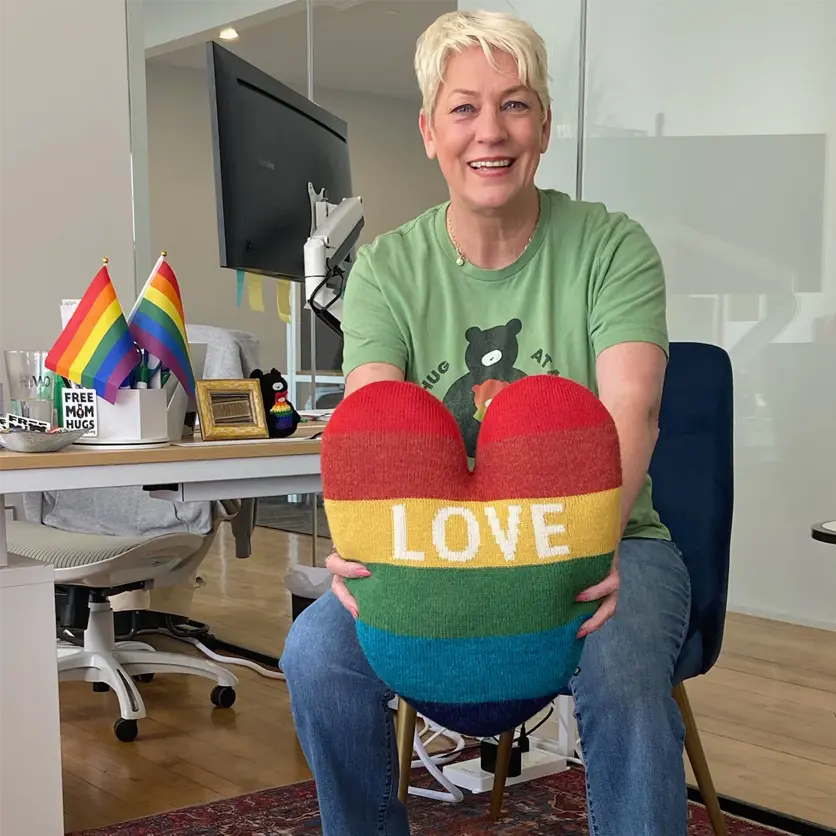 Person smiling and holding a heart-shaped rainbow pillow with LOVE text, seated in an office with rainbow flags.