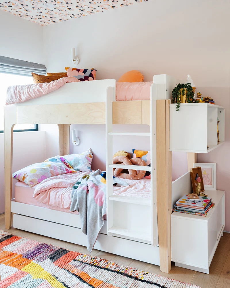 A brightly decorated childrens room with a wooden bunk bed, colorful bedding, shelves holding toys and books, and a patterned ceiling and rug.