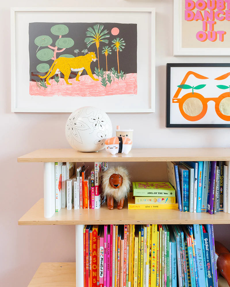 Bookshelf with colorful childrens books, a plush lion, a white decorative ball, and framed art on the wall, including a leopard in a jungle scene and a motivational quote.