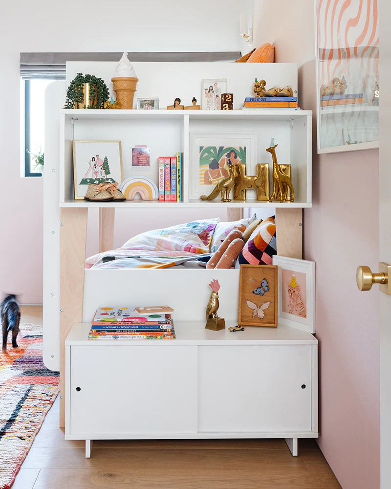 Childrens bedroom with a loft bed surrounded by shelves holding books, toys, and decor items. A colorful rug is on the floor, and a partially open door reveals a pet entering the room.