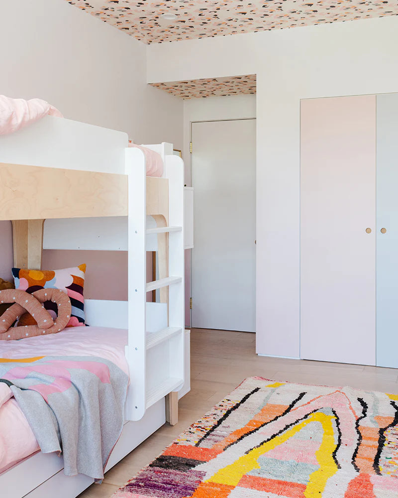Childrens room with a white bunk bed, colorful bedding, and a vibrant geometric rug. Walls and ceiling have a multicolored pattern. White wardrobe and a closed door are visible.