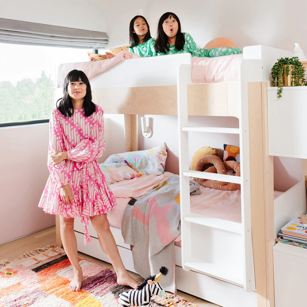 A woman in a pink dress stands beside a bunk bed. Two children in green pajamas sit on the top bunk. Decor includes stuffed animals and a small plant. A colorful rug is on the floor.