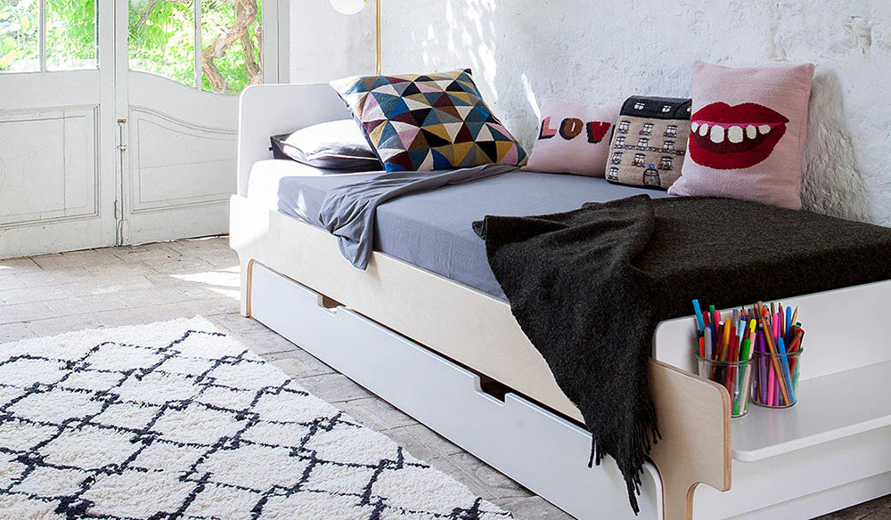 A cozy daybed with a gray mattress and colorful pillows against a white brick wall. A black blanket is draped over the bed, and colored pencils are in a holder by the side.