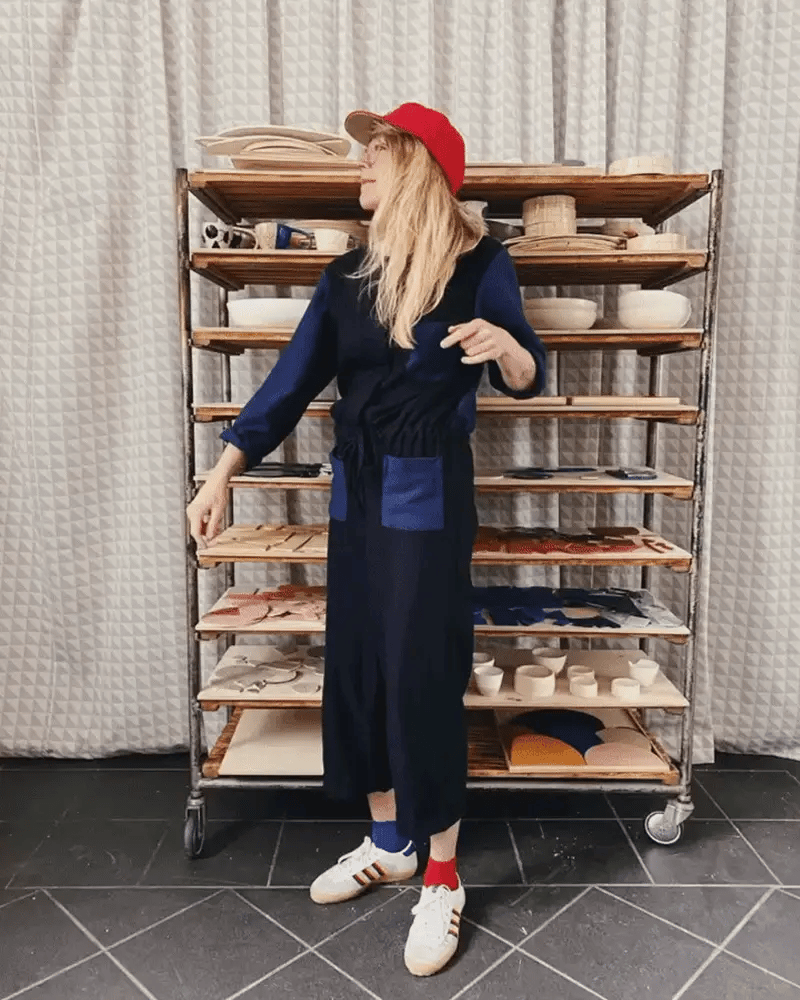 Person in a red cap and dark apron standing in front of a wheeled rack with pottery shelves, holding a pottery tool, with a gray curtain backdrop.