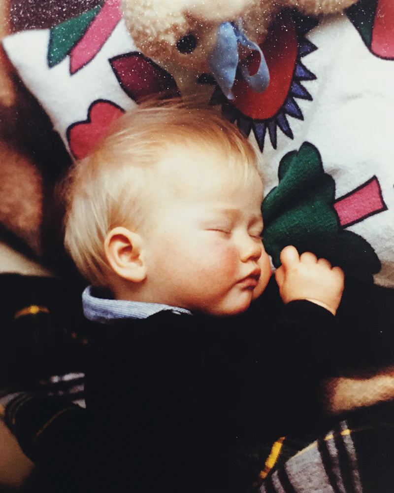 Baby sleeping peacefully on a patterned pillow, holding a green plush toy, with teddy bear in the background.