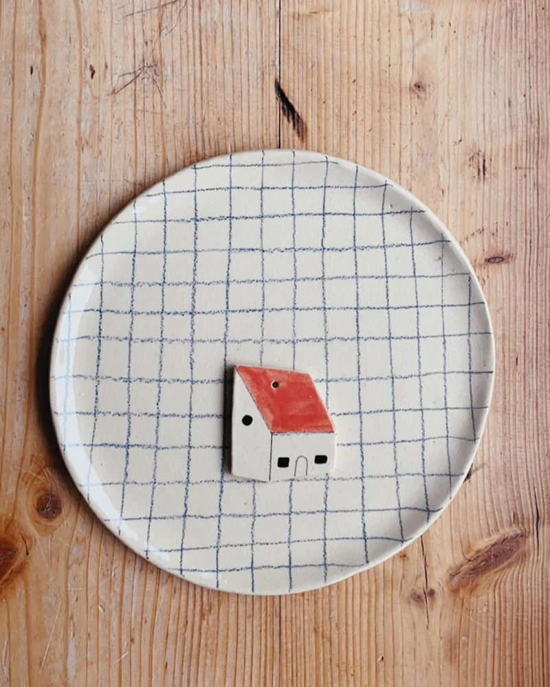 Ceramic plate with a grid pattern, featuring a small house-shaped ornament with a red roof and two black windows, on a wooden surface.