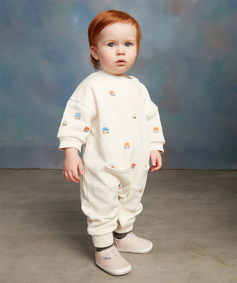 A toddler with red hair stands in a neutral studio setting, wearing a cream-colored onesie and pink shoes.