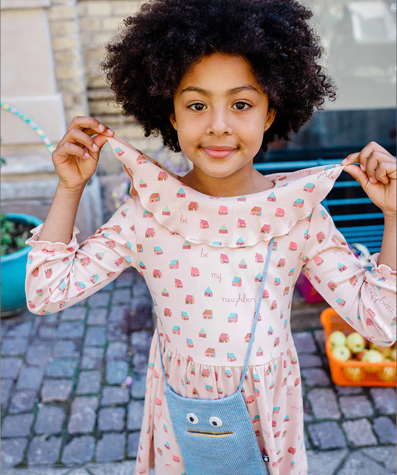 A young girl with curly hair holds her collar, wearing a patterned dress and a blue crossbody bag, standing on a cobblestone street.