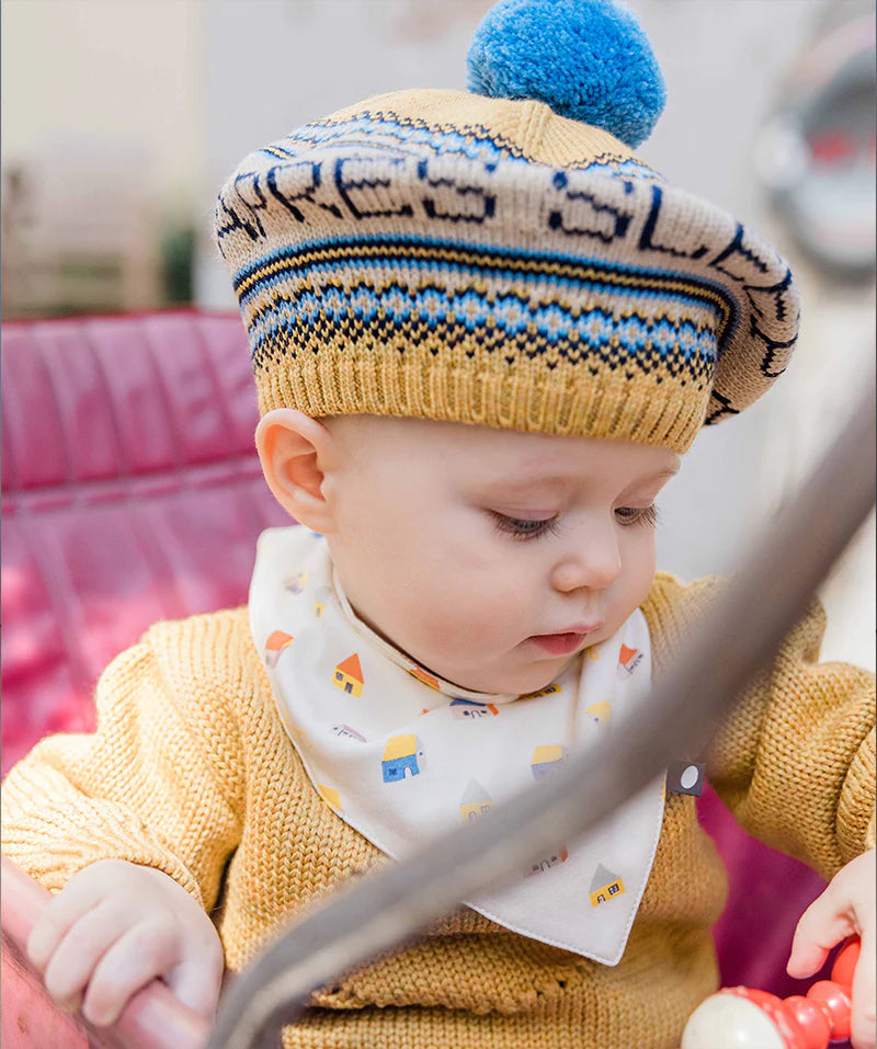 Baby wearing a patterned knit hat and a bib with house designs, sitting in a red seat, looking down.