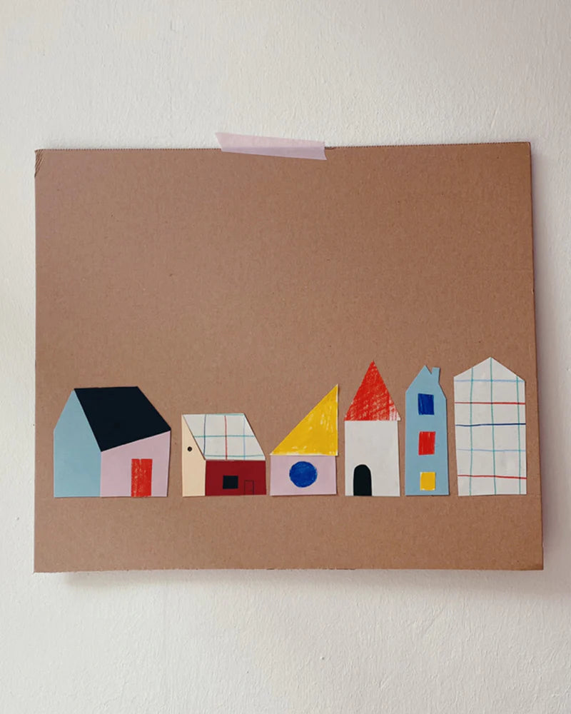 A cardboard sheet featuring colorful paper cutouts of houses in various simple shapes and sizes, taped to a white wall.