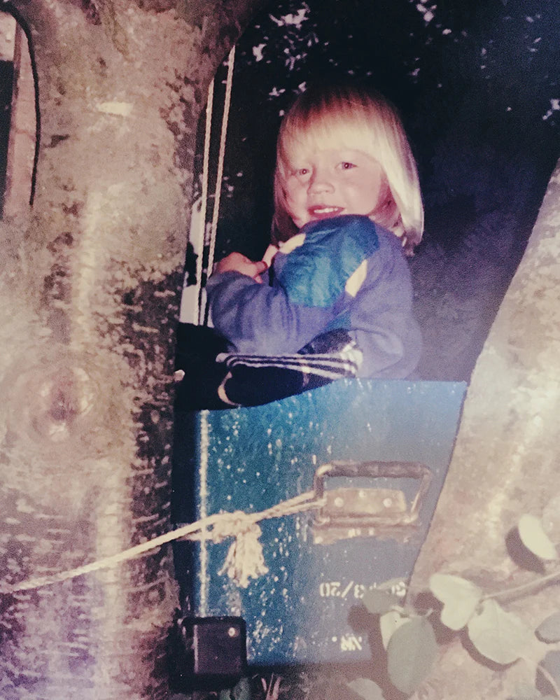 Child with blonde hair sitting in a treehouse, holding a blue item, surrounded by tree branches.