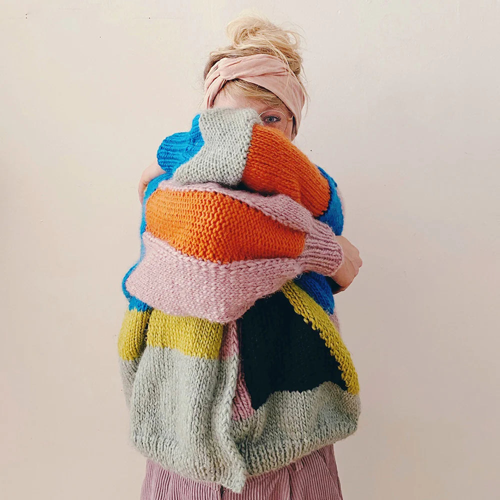 Person holds a large, colorful knitted blanket, covering most of their face. They wear a pink headband and have blonde hair.