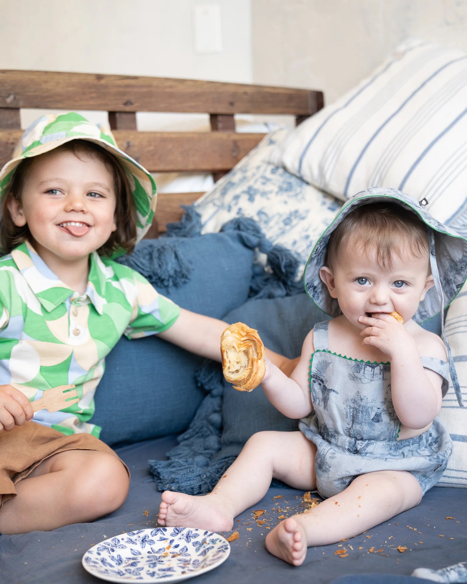 Two young children sitting on a sofa with blue and white pillows. The older child wears a green hat and shirt, the younger child wears a blue romper and hat, holding a pastry.